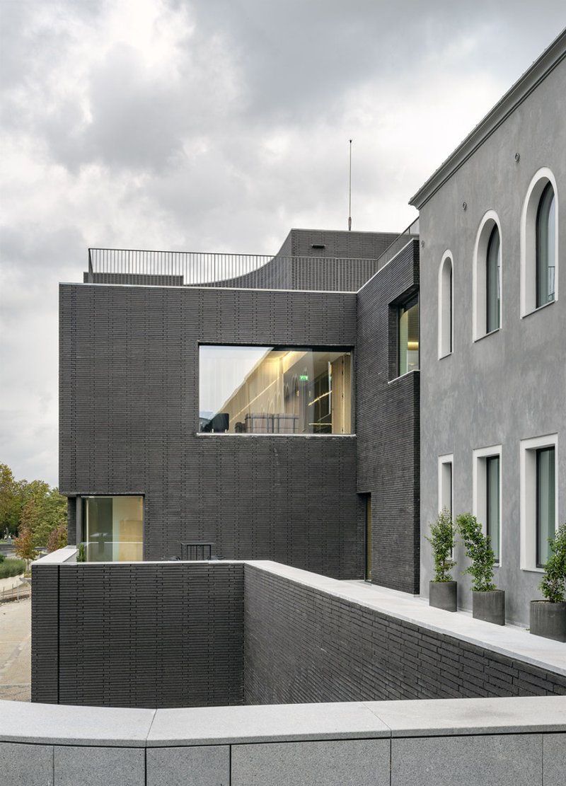 Dark brick addition framing a window into the lobby beside an existing grey plaster facade under cloudy skies