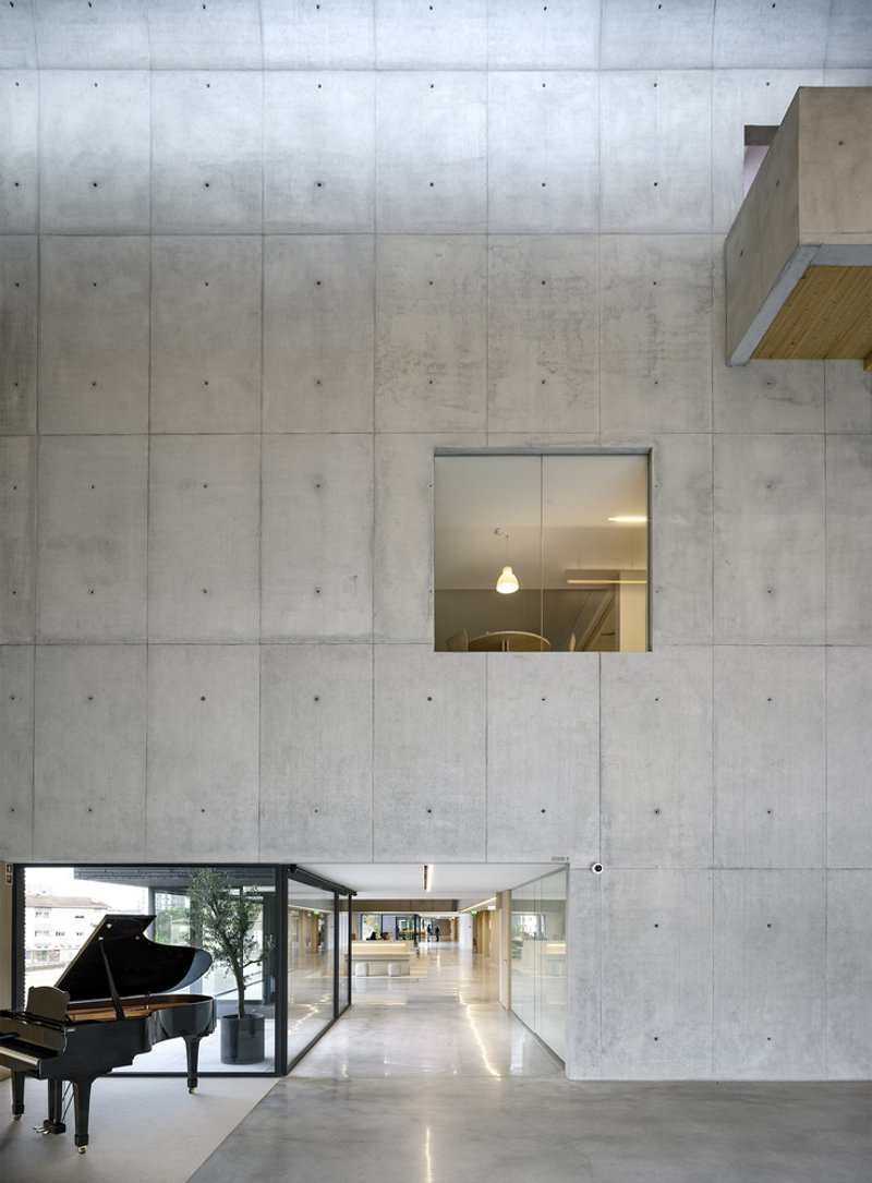 Double-height lobby with board-formed concrete walls and a grand piano visible through glass doors at dusk