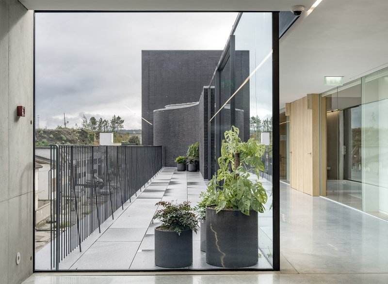 View through glass doors to rooftop terrace with dark brick volumes and planters under cloudy sky