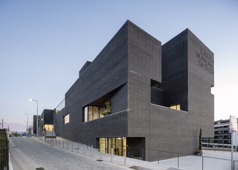 Dark brick facade with recessed windows and illuminated openings at twilight along sloped street