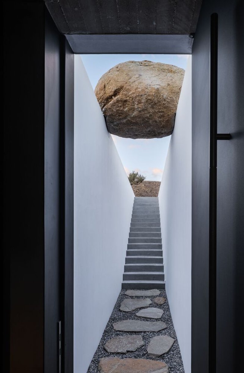 Courtyard corridor with stone paver path leading to stairs beneath suspended boulder and sky