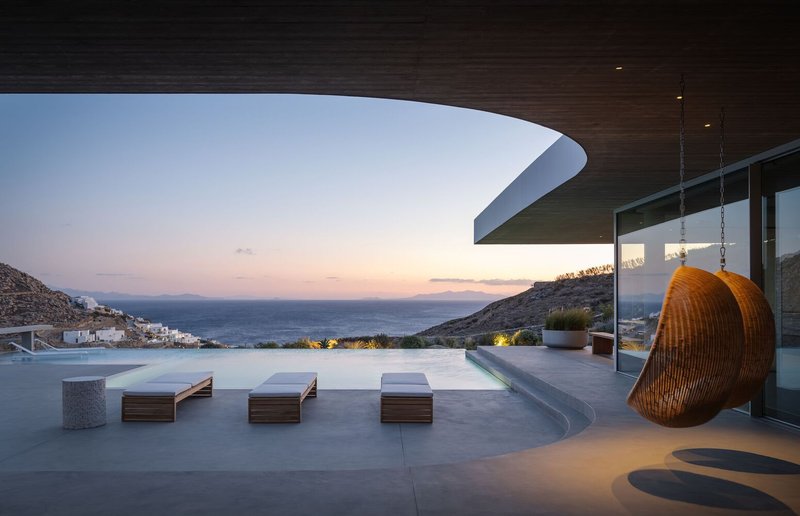 Poolside terrace with woven hanging chair and curved roof overhang at dusk