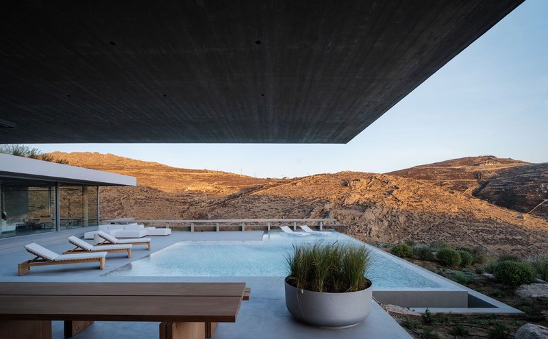 Infinity pool terrace with timber loungers beneath a cantilevered soffit facing rocky hills at sunset