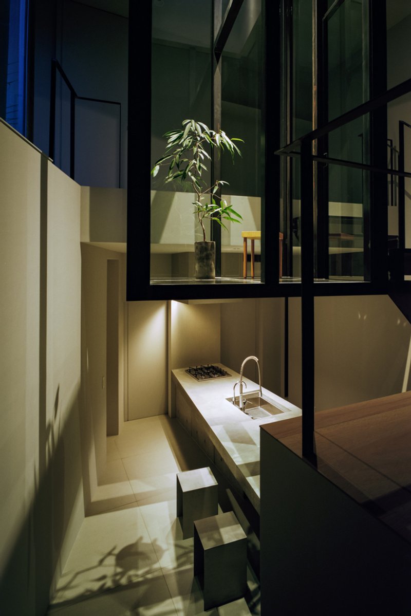 Night view through steel-framed glazing showing potted plant and kitchen counter with warm interior lighting
