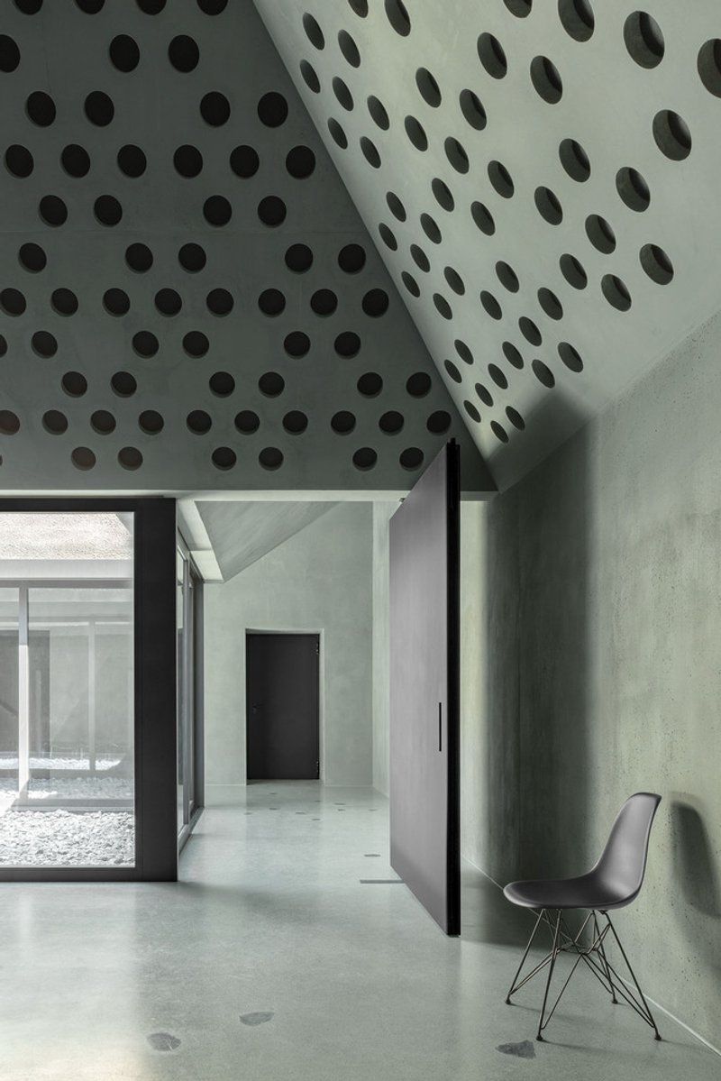 Perforated concrete ceiling with circular openings above a doorway and single chair against green wall