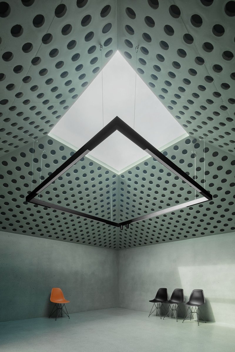 Looking up at pyramidal skylight framed by perforated concrete ceiling with chairs below