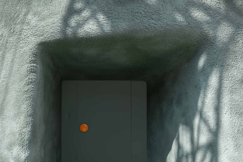 Textured stucco corridor with dappled shadows leading to grey door with orange circular handle