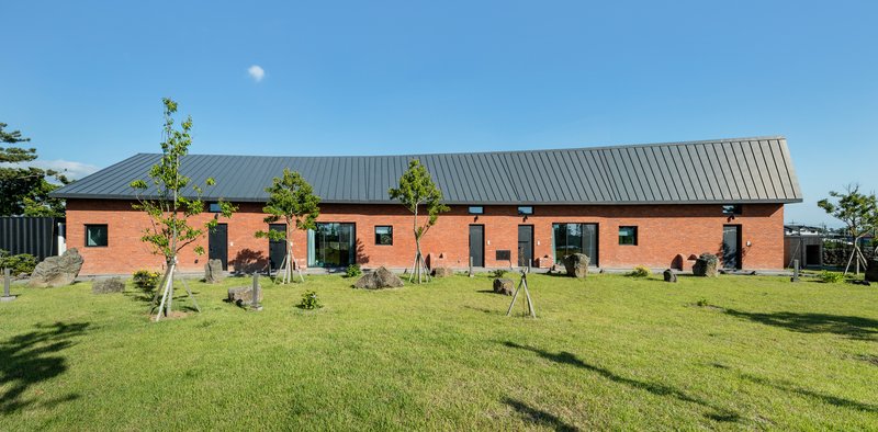 Red brick facade with standing seam metal roof surrounded by young trees and lawn in clear daylight