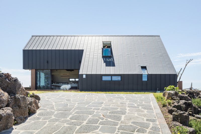 Angular metal-clad volume with steeply pitched roof and stone paving terrace against a blue sky