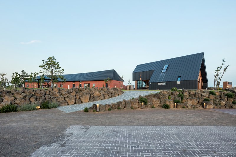 Paved entry plaza with volcanic rock retaining wall leading to clustered gabled volumes