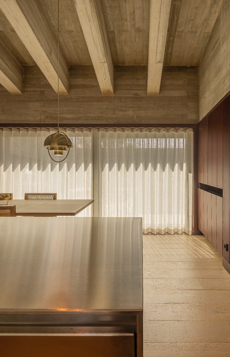 View of the stainless steel island and backlit sheer curtains beneath a timber plank ceiling with structural beams