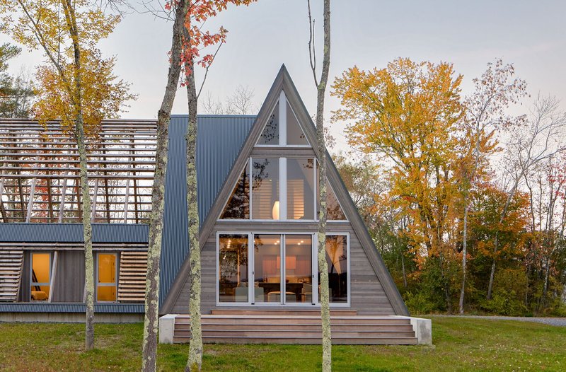 Triangular gabled volume with horizontal wood siding and full-height glazing among autumn trees at dusk