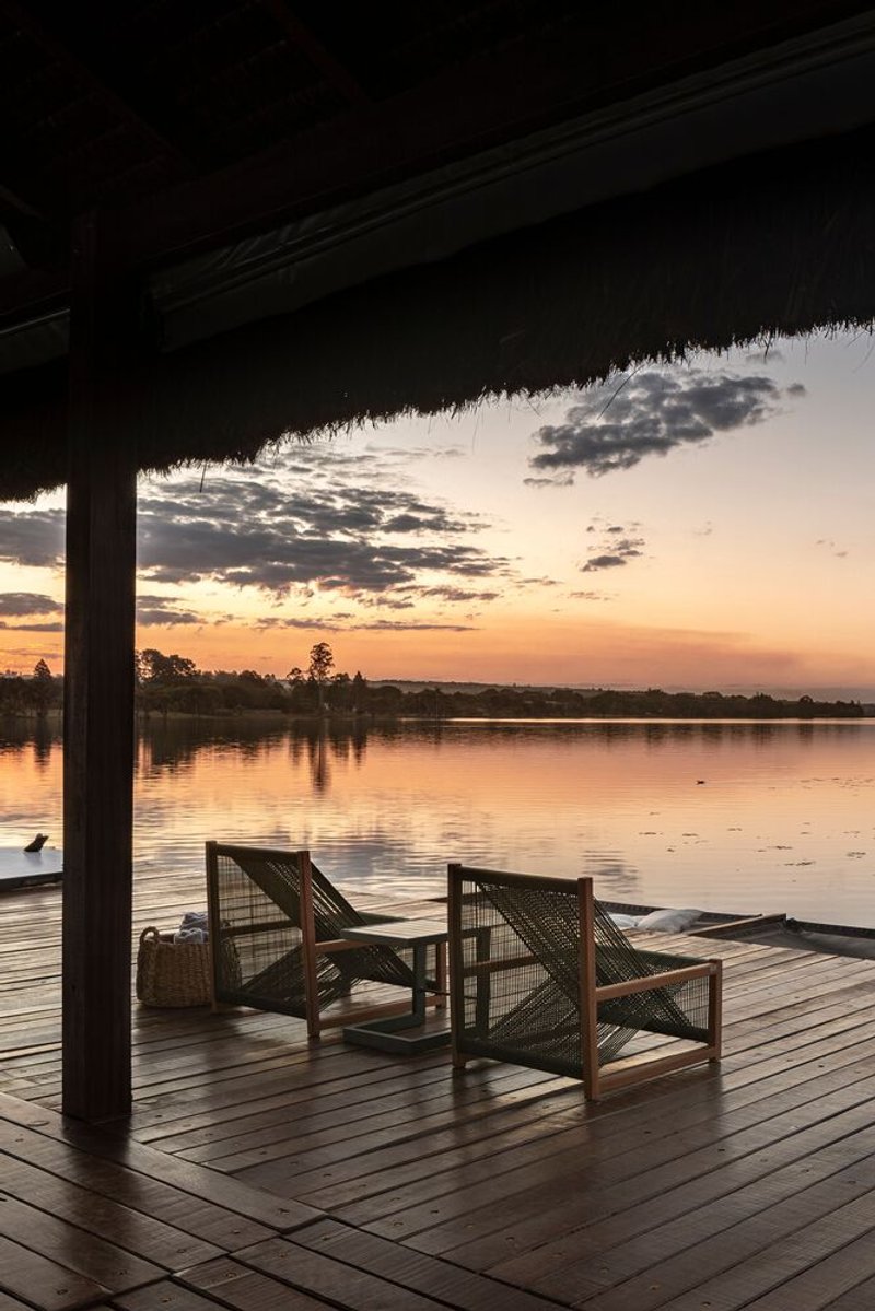 Timber deck with woven lounge chairs overlooking still lake at sunset beneath thatched roof overhang