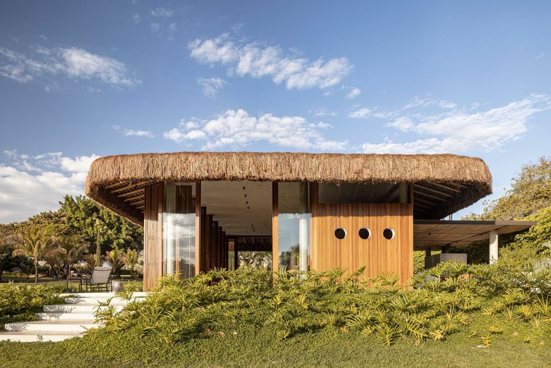 Thatched roof pavilion with timber and vertical wood cladding set within a planted lawn under afternoon clouds