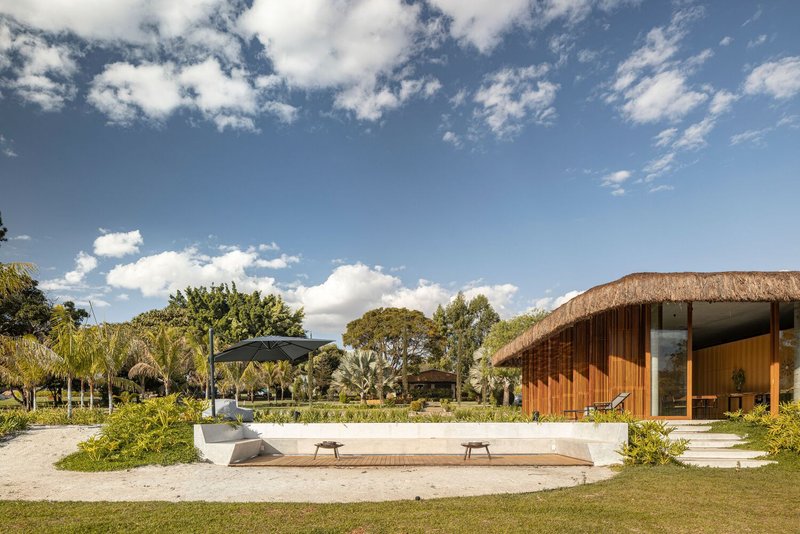 Timber pavilion with thatched roof beside a paved terrace and lawn under scattered clouds