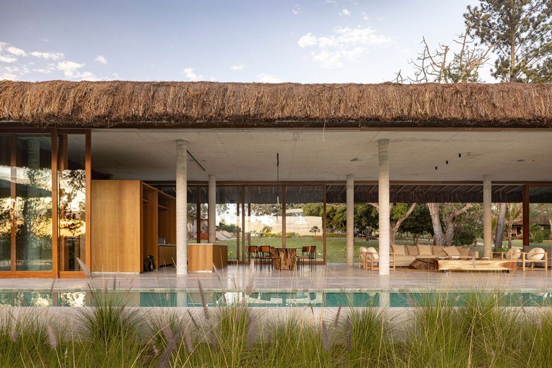 Open-air pavilion with concrete ceiling, white columns and thatched roof beside reflecting pool and grasses
