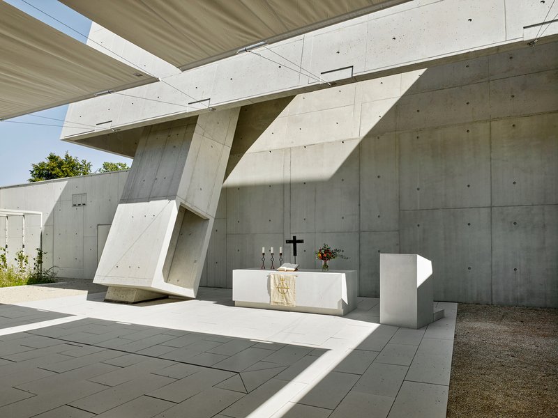 Outdoor altar beneath angled concrete canopy supported by faceted columns in summer sunlight
