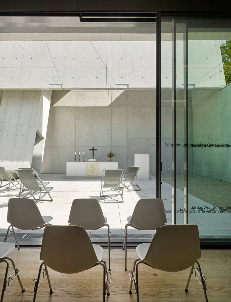 View through glass doors from interior seating toward outdoor altar framed by concrete walls and angled structure