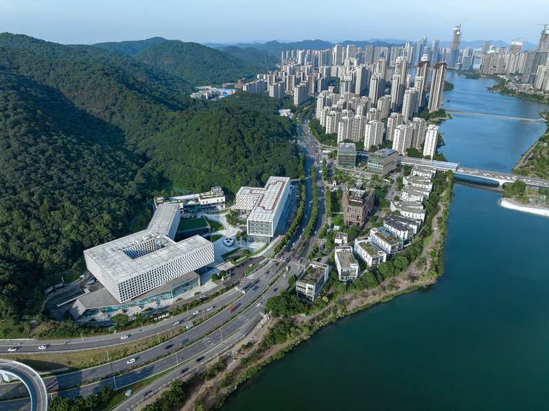 Drone view showing the complex positioned between forested hills and a curving river with highway infrastructure