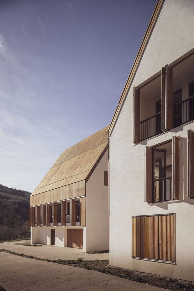 Two gabled volumes with white plaster walls and vertical timber shutters on a sloped paved street