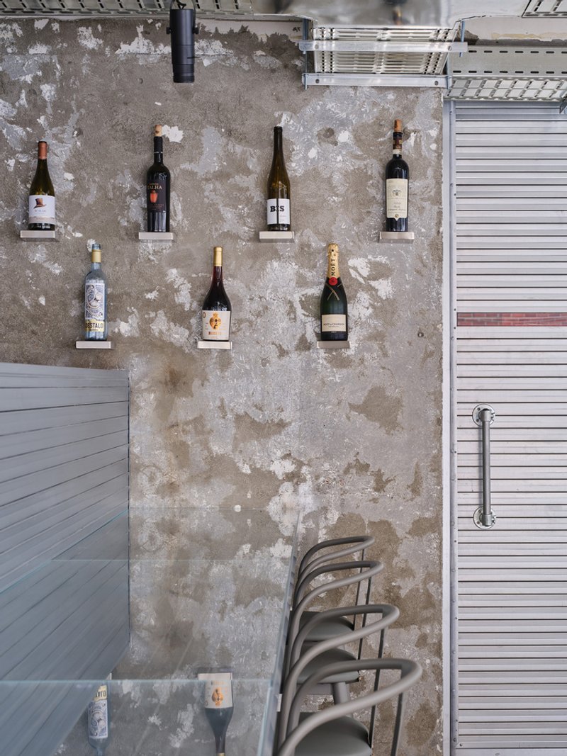 Weathered concrete wall displaying wine bottles on mounted shelves with stacked chairs below