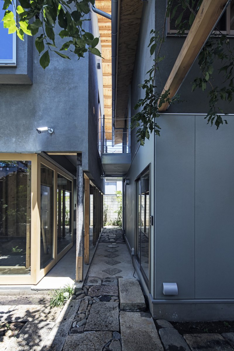 Narrow paved passage between plaster and panel walls with exposed rafters and overhanging tree branches