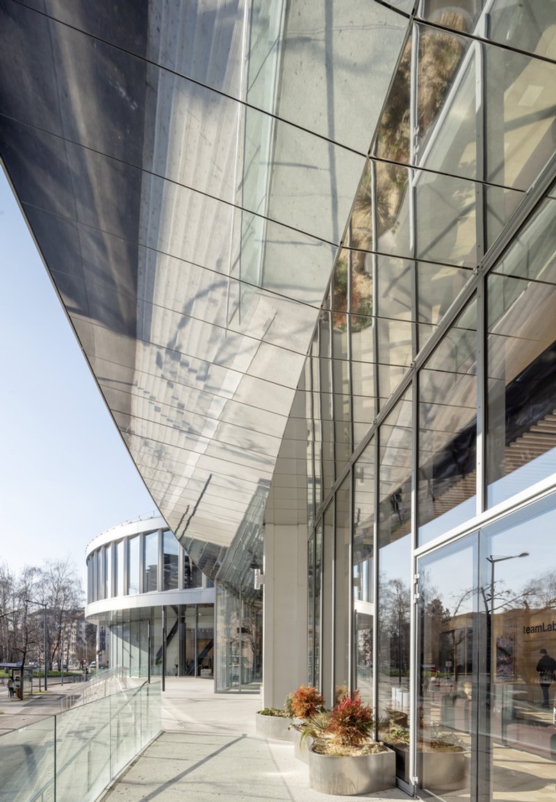 Ground-level view of the glass curtain wall entrance with overhead canopy reflecting bare trees