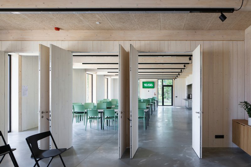 View through open double doors into a cafeteria with rows of green chairs under exposed timber ceiling beams