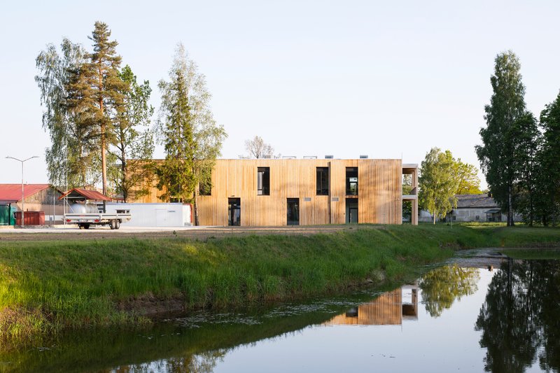 Timber-clad building reflected in a pond at dusk with birch trees and a red warehouse in the background
