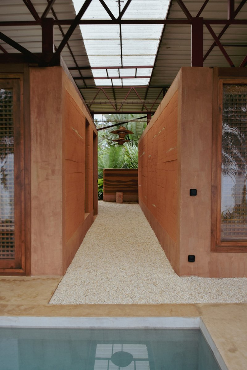 Gravel corridor flanked by rammed earth walls with exposed steel truss roof above a pool edge