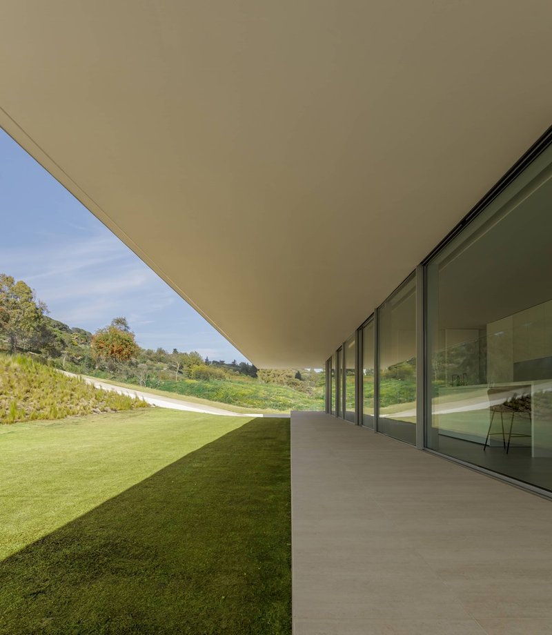 Covered terrace with continuous glazing running parallel to a lawn sloping up the hillside