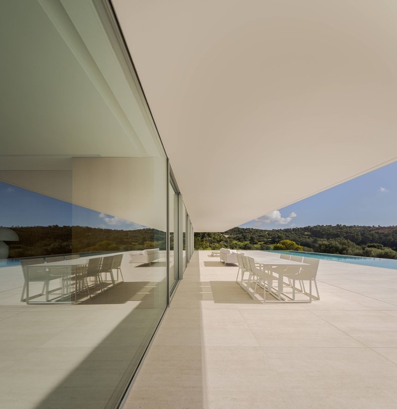 Outdoor terrace with white dining furniture beneath a deep overhang and reflected in the glazing