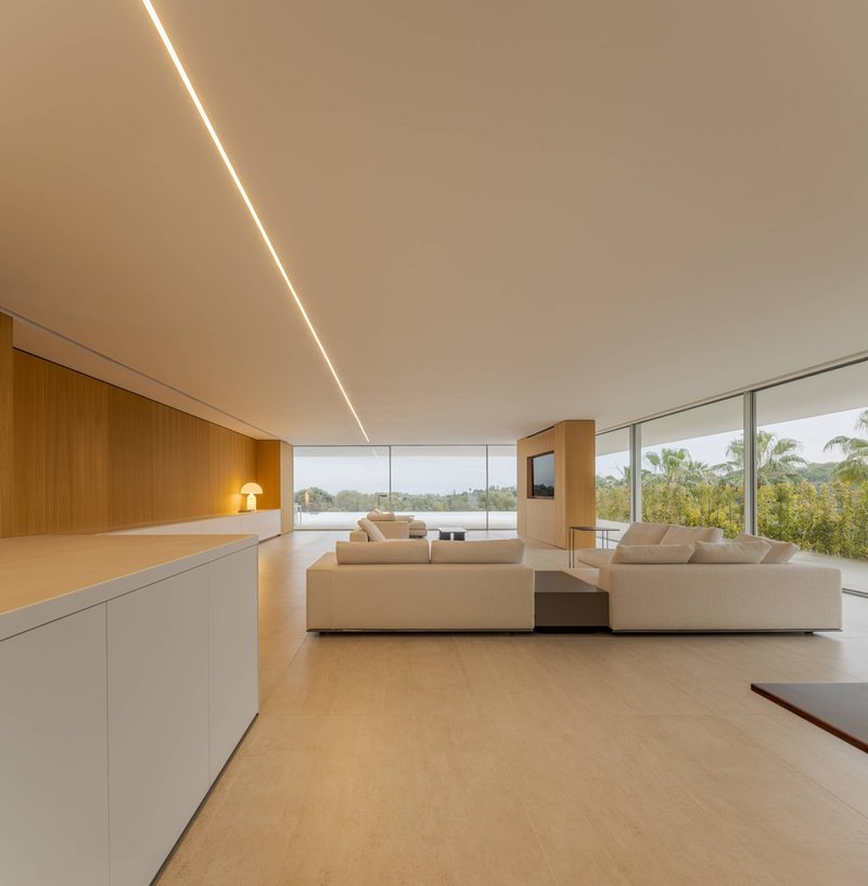 Open living room with continuous ceiling light slot and floor-to-ceiling glazing overlooking tropical landscape