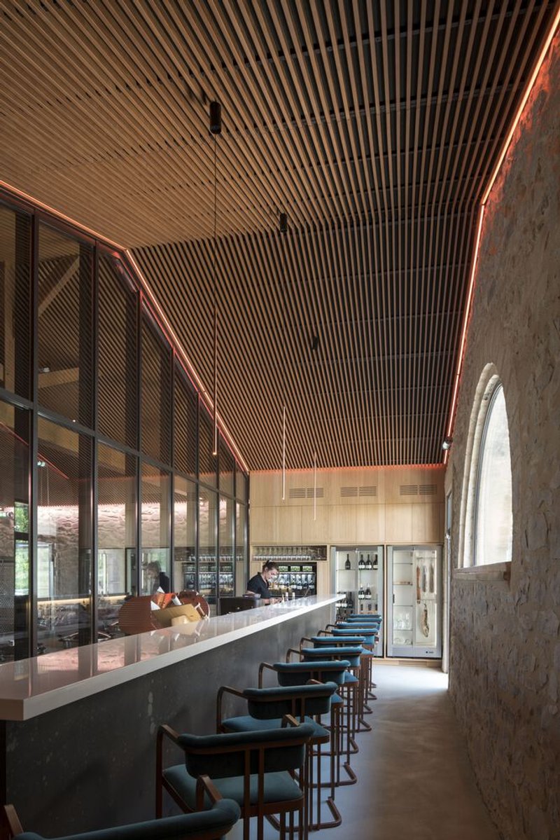 Bar seating area with timber slat ceiling, glazed partition wall, and arched stone wall with natural daylight