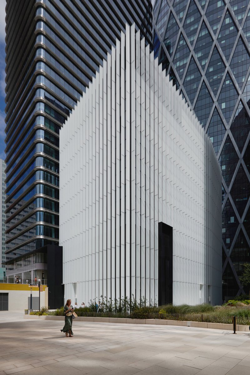 White vertically ribbed facade set between two high-rise towers as a pedestrian passes on the plaza