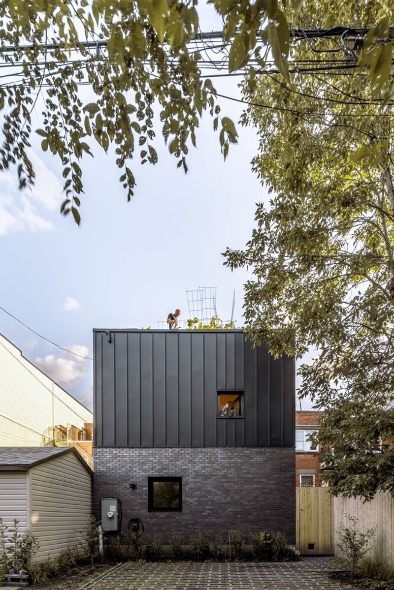 Street view of the two-story volume with dark metal cladding over brick base beneath overhanging birch branches