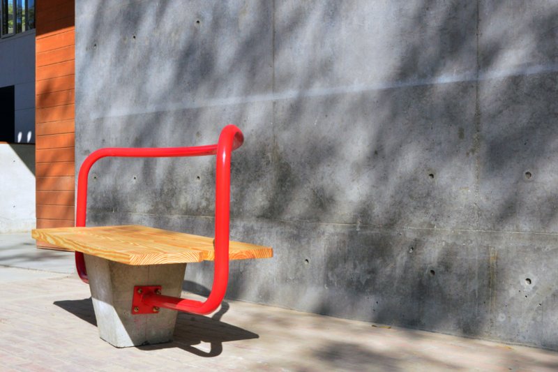 Outdoor bench with timber seat and red tubular steel frame against a weathered concrete wall