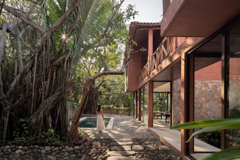 Covered walkway with timber columns and stone walls alongside a pool and banyan tree roots
