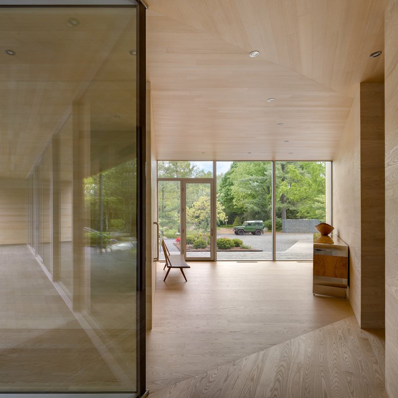 Wood-lined corridor with glass partition reflecting view through glazed doors to landscaped driveway