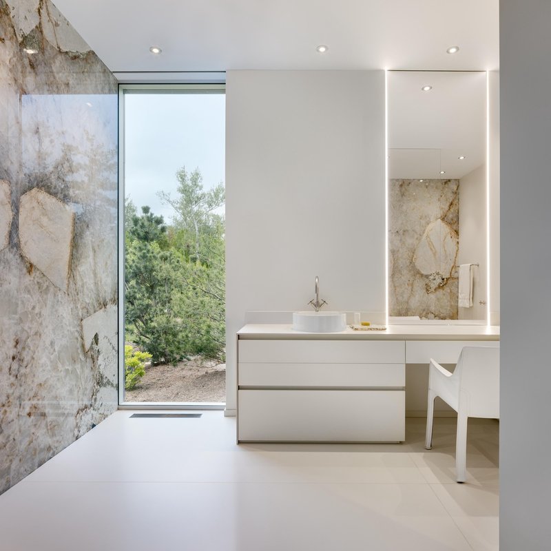 Bathroom with weathered stone wall, floor-to-ceiling glazing overlooking green landscape and white vanity with backlit mirror
