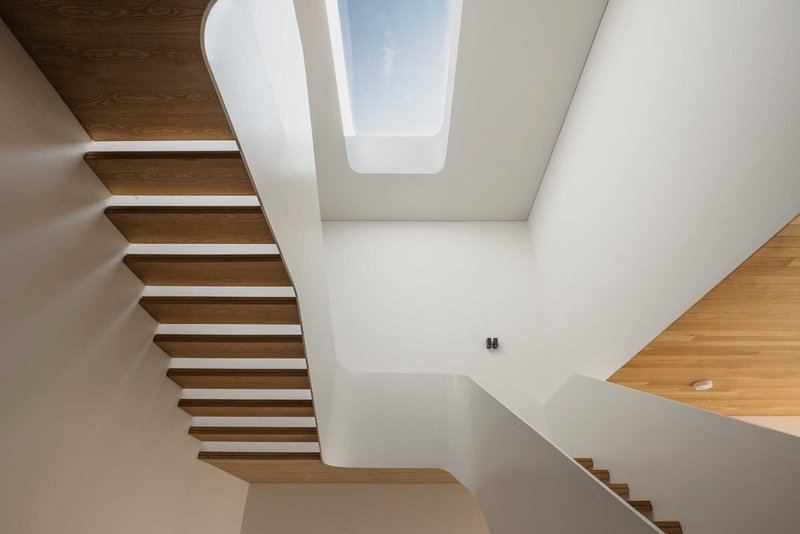 View up the cantilevered timber stairwell toward a rectangular skylight framed by white walls