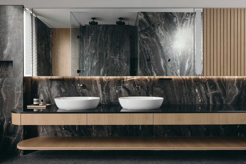 Double vanity with vessel sinks on timber cabinetry below a backlit mirror and dark marble walls