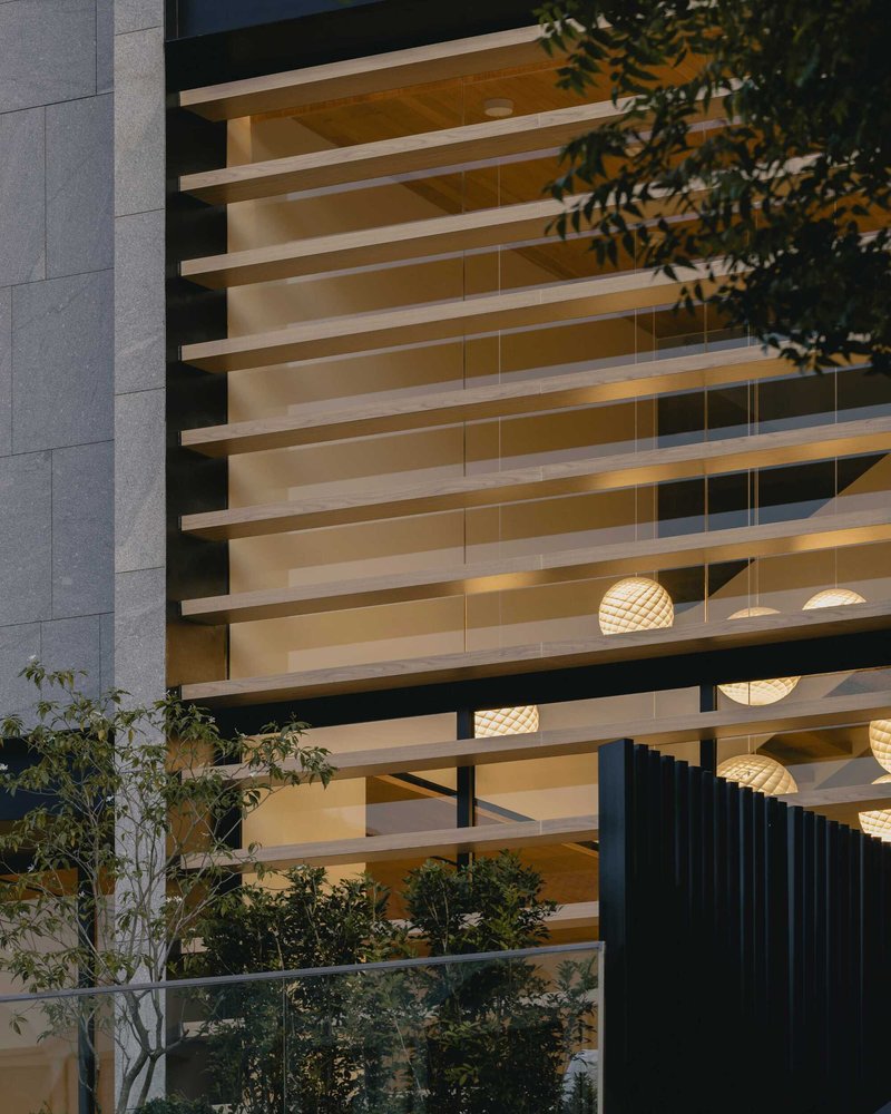 Facade detail showing horizontal timber louvers and pendant lights visible through the glazing at dusk