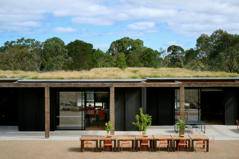 Long elevation of the dark building with its green roof of native grasses and a row of timber dining tables at the foreground