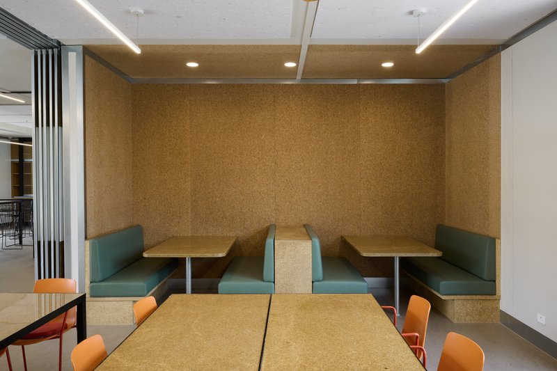 Interior booth seating nook with cork walls, teal banquettes and cork-topped tables under linear lights