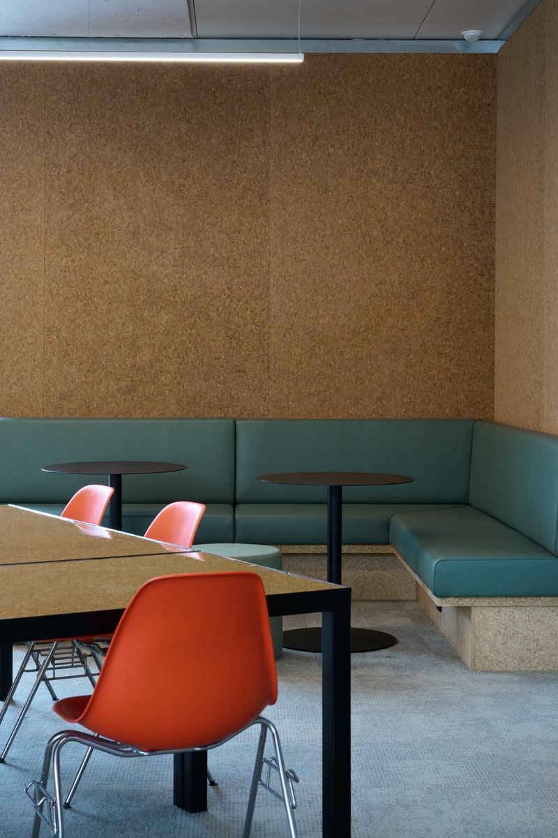 Interior breakout nook with a cork acoustic wall, teal banquette seating, and orange moulded chairs at cork-topped tables