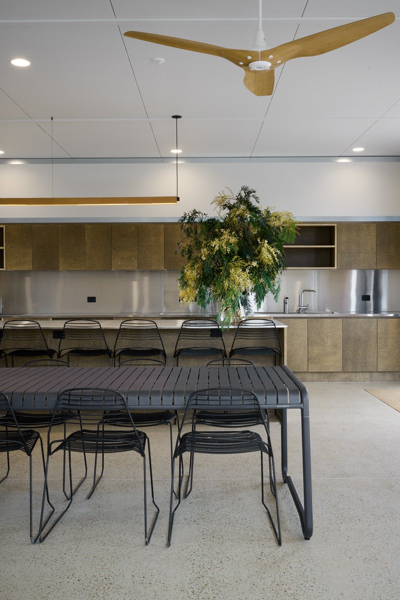 Communal kitchen and dining space with a long table, wire chairs, timber cabinetry and a tall indigenous foliage arrangement