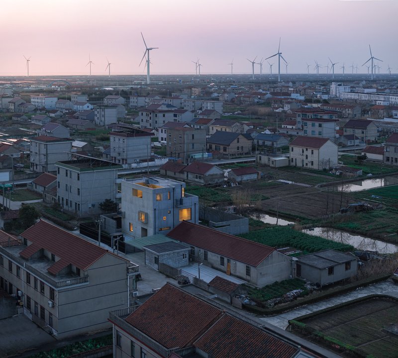 Aerial view of illuminated concrete house among low-rise village buildings with wind turbines on the horizon
