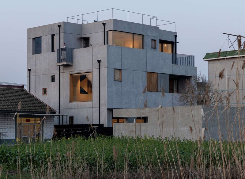 Stacked concrete volumes with illuminated openings at dusk above a grassy foreground with dry reeds