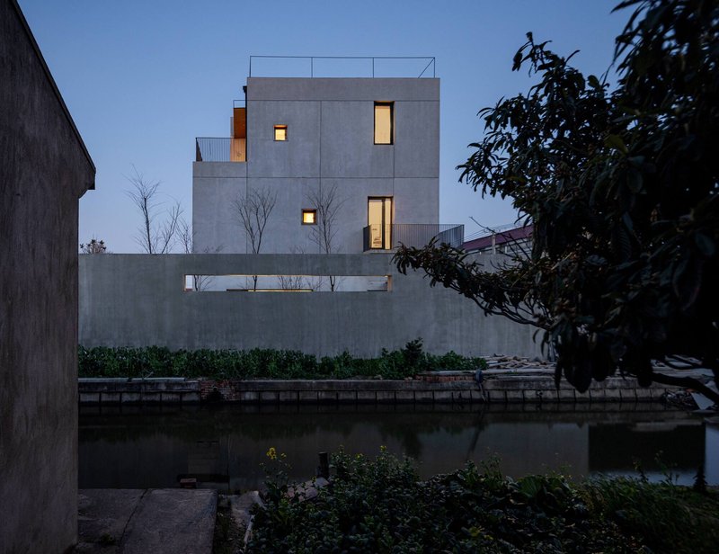 Street facade with punched window openings and bare trees above a canal at dusk